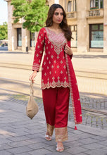 Load image into Gallery viewer, Woman in a red traditional outfit with gold embroidery standing on a street.
